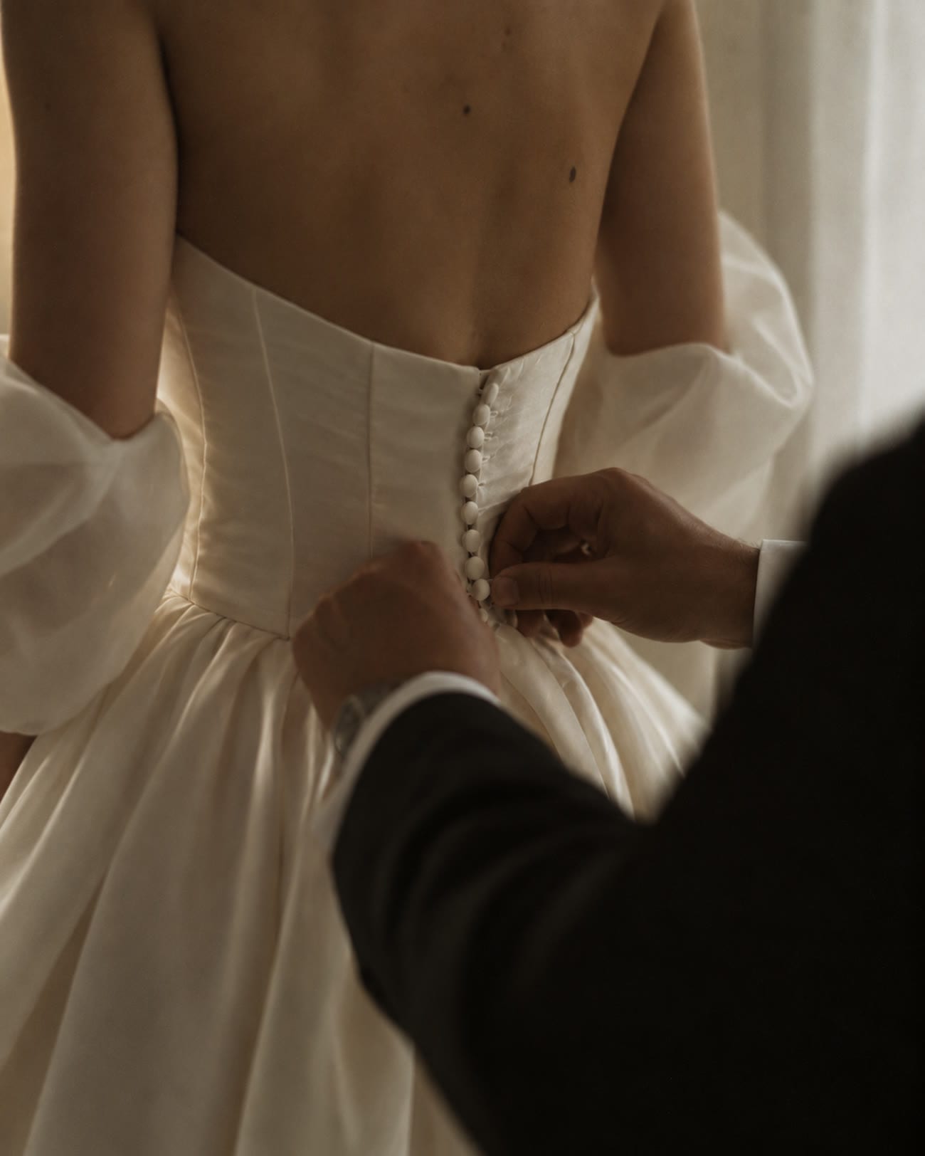 A bride's dress being buttoned by hand in soft window light