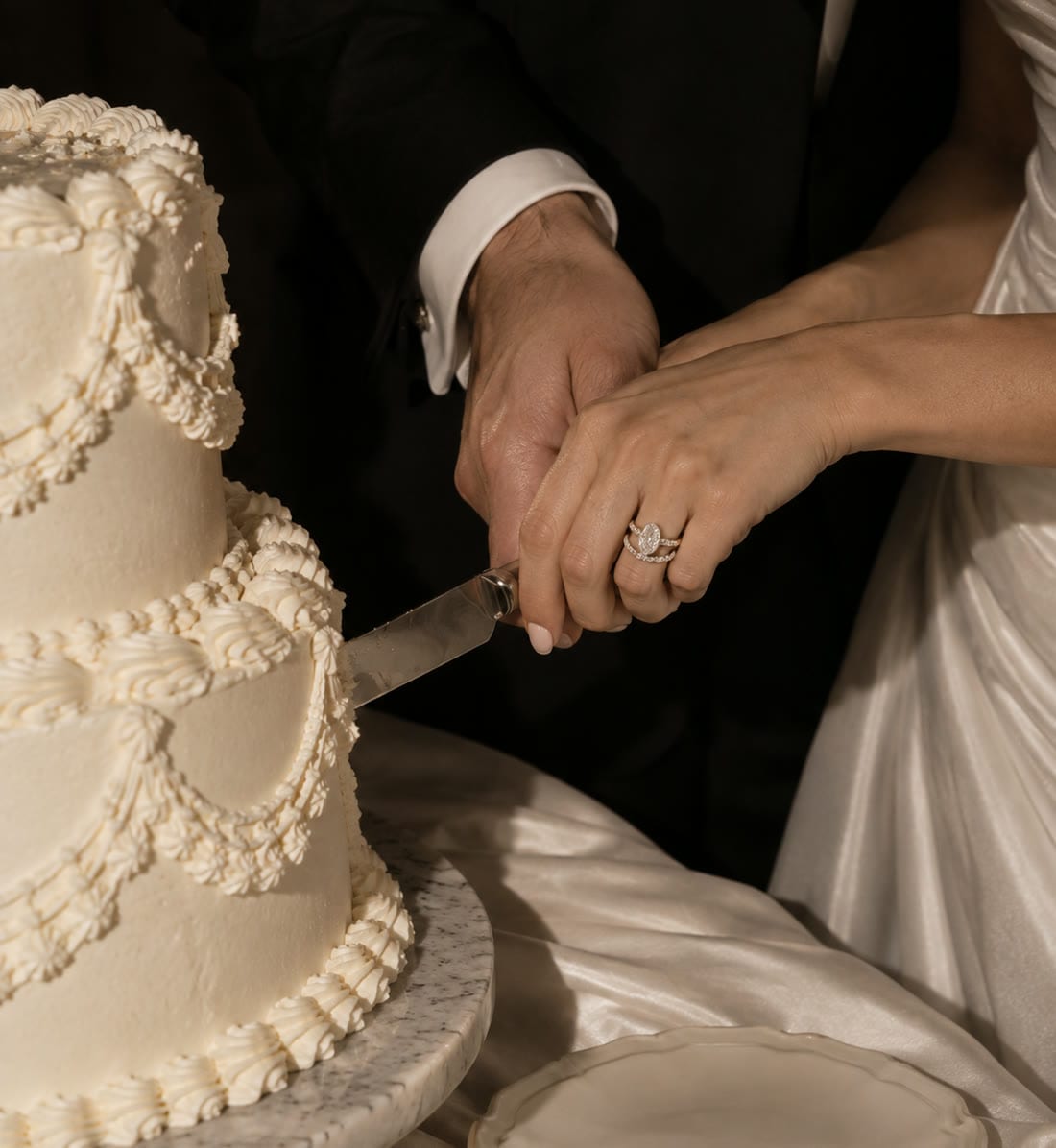 A couple's hands together on the knife, cutting a fluted ivory wedding cake